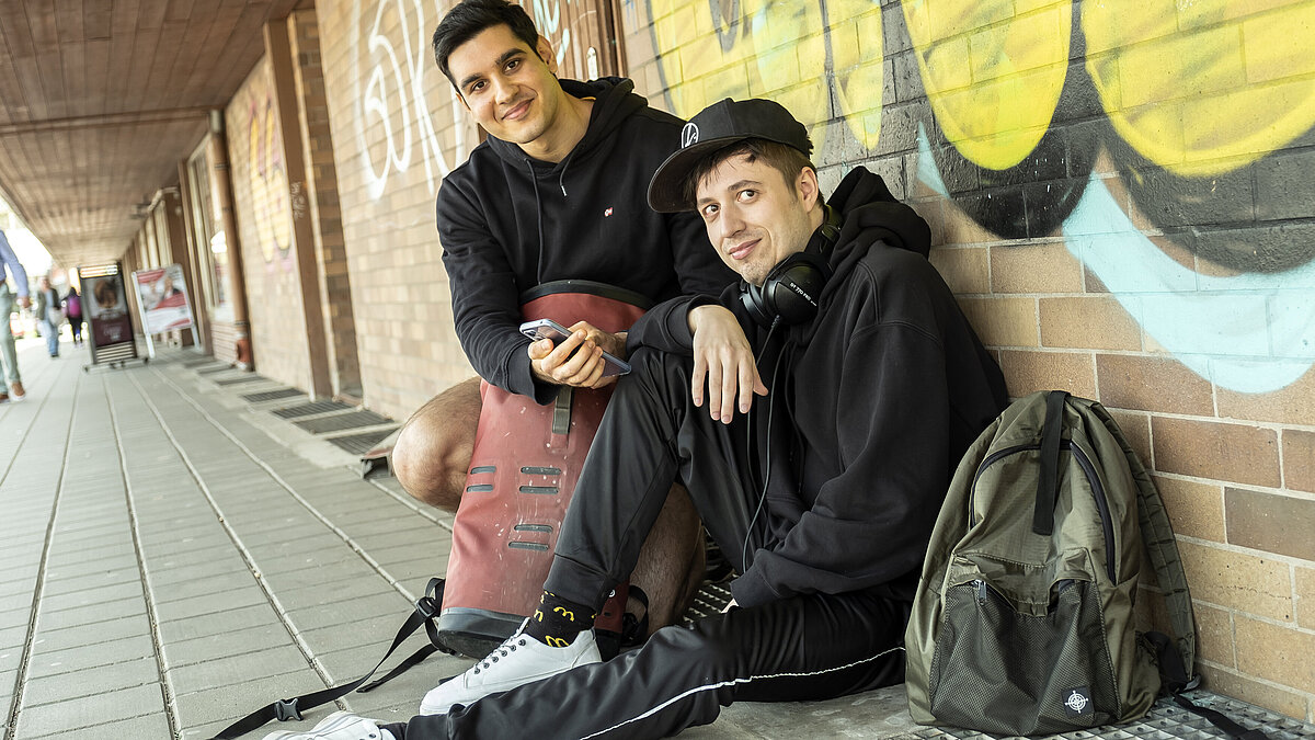 Zwei junge Männer sitzen mit Rucksäcken und Handy vor einer Wand.